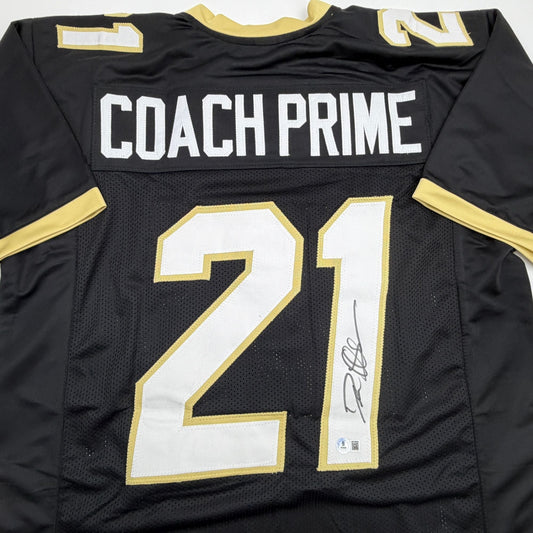 Back view of black college football jersey with gold and white trim, "COACH PRIME" text, number 21, and signature.