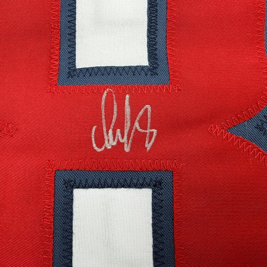 Close-up of white autograph signature on red jersey fabric with dark navy and white trim framing.