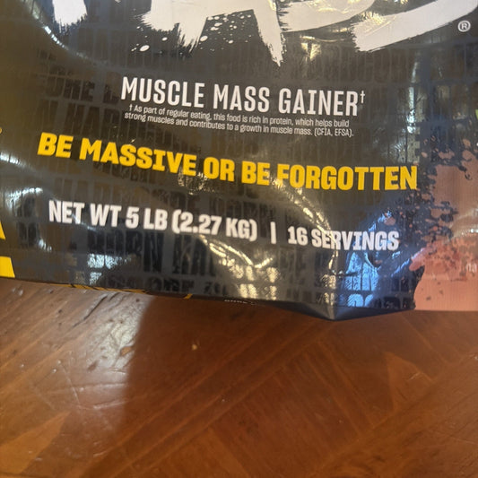 Close-up of black Mutant Mass 5LB container showing "Muscle Mass Gainer" text and yellow "Be Massive or Be Forgotten" slogan on wooden surface.