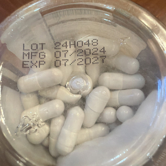 Close-up of white capsules in clear plastic container with printed lot number and expiration date visible on lid.