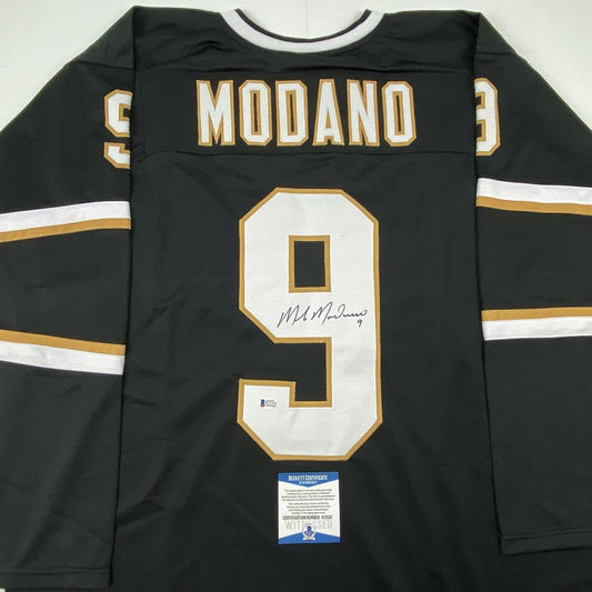Back view of black Dallas hockey jersey number 9 with "MODANO" nameplate, gold and white trim, signed in blue ink, Beckett authenticity certificate visible at bottom.