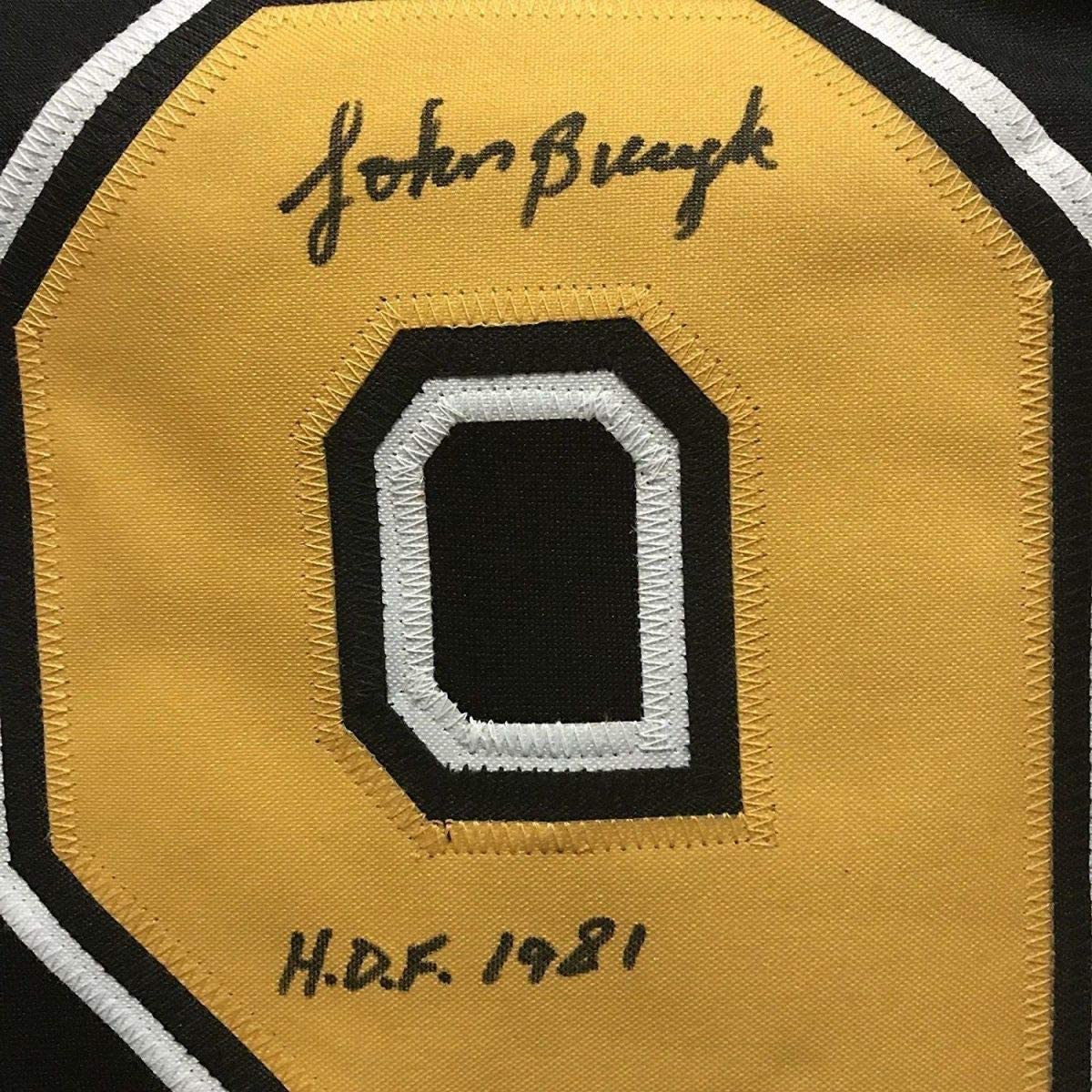 Close-up of signed yellow hockey jersey patch showing number 0 with white and black trim, signed "John Bucyk" at top and "H.D.F. 1981" at bottom.