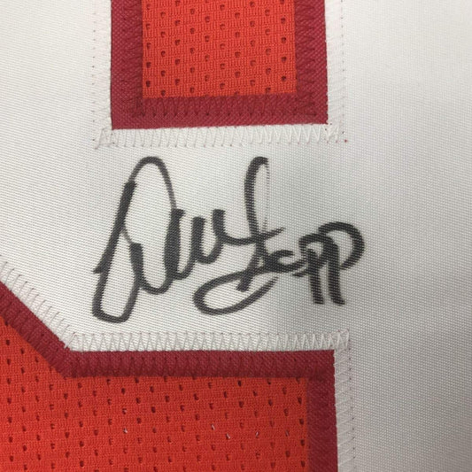 Close-up of a white jersey panel with a black ink autograph, bordered by red stitching and framing orange mesh numerals.