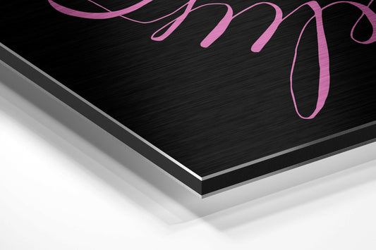 Angled perspective view of black brushed aluminum dibond panel with pink cursive "Believe" lettering, showing layered edge and white base surface below.