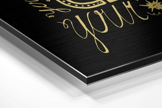 Angled close-up of black brushed aluminum wall art with gold cursive lettering and decorative elements, showing layered edge detail.