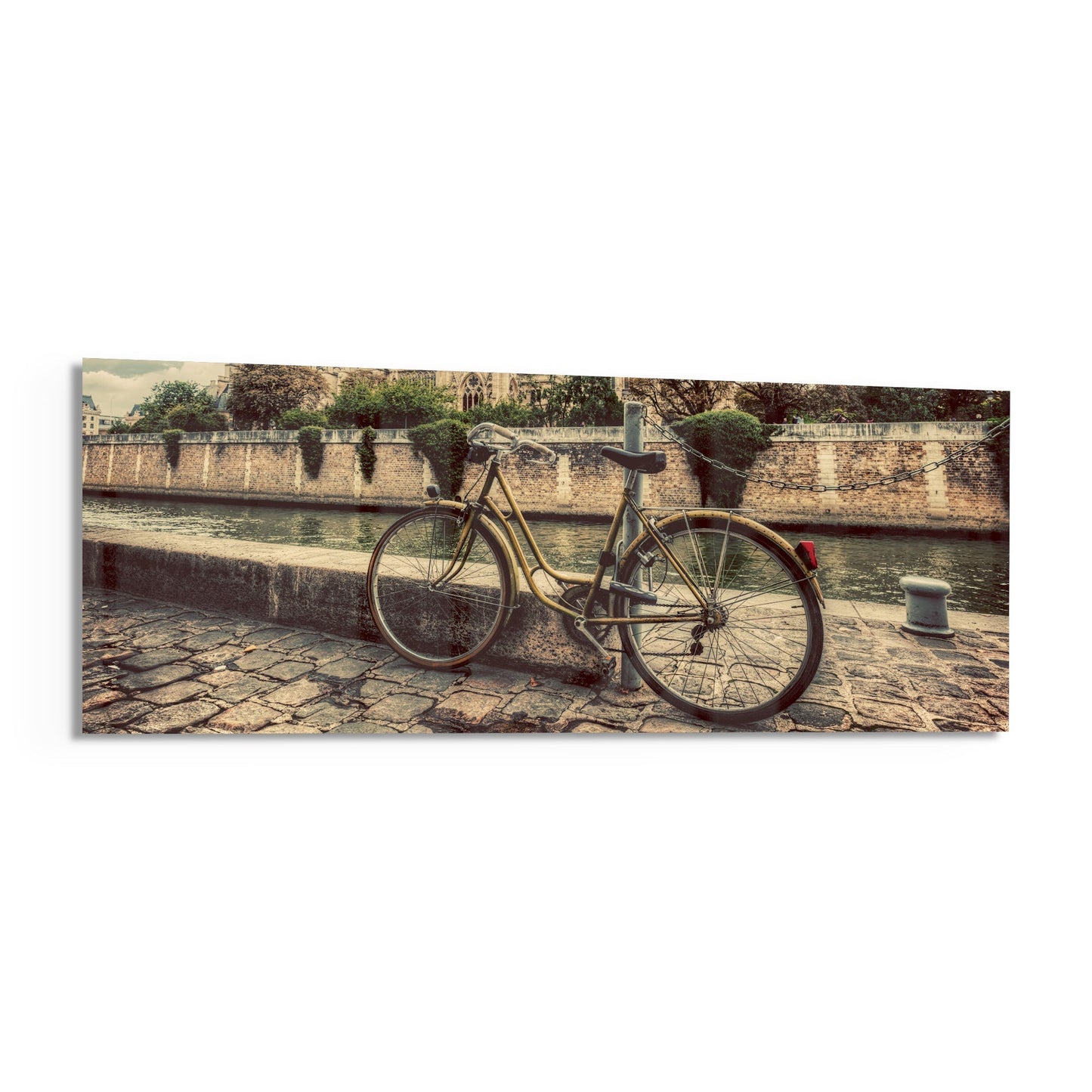 Vintage bicycle with red rear light leaned against stone wall beside calm river, with historic fortification and green trees in background.
