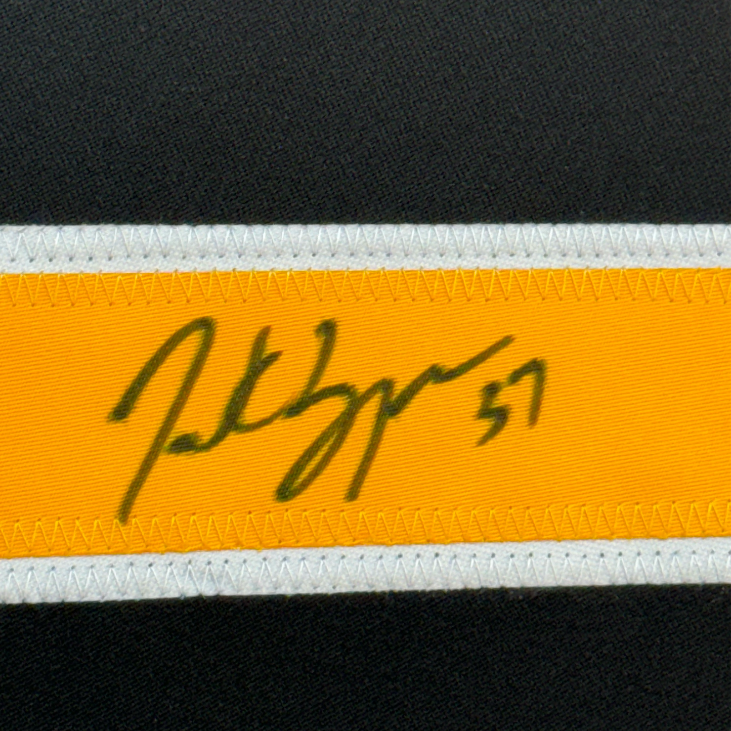 Close-up of handwritten autograph on yellow ribbon strip with white borders against dark fabric background.