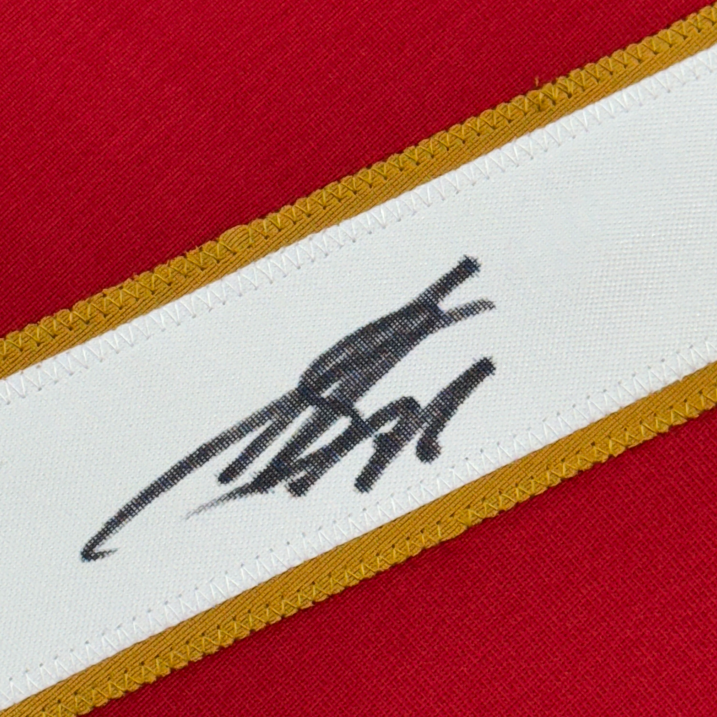 Close-up of autographed signature in black ink on white fabric stripe with red background and gold trim.