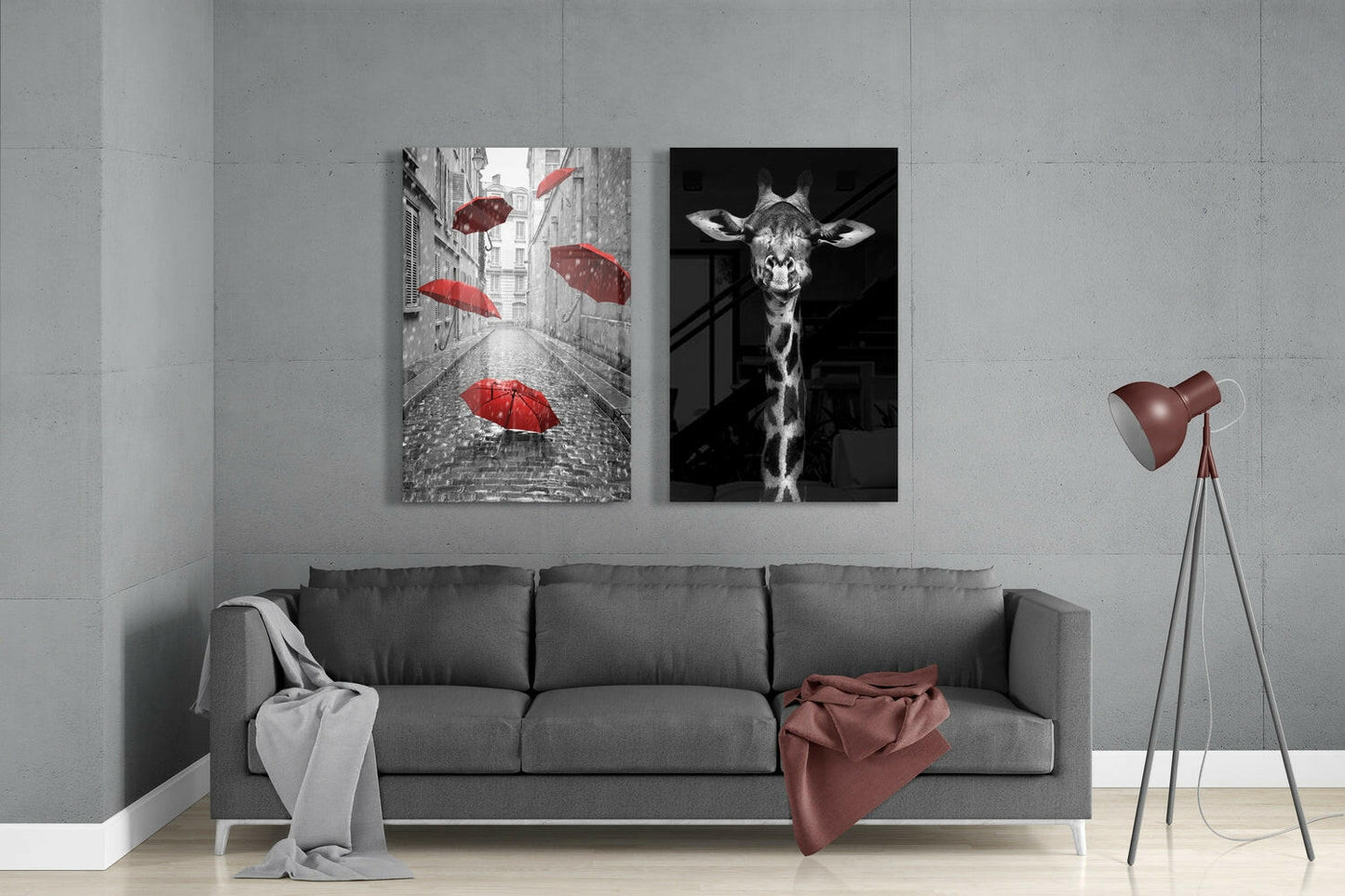 Modern living room with gray sofa displaying two glass wall art panels: red umbrellas on cobblestone street and black-and-white giraffe portrait.