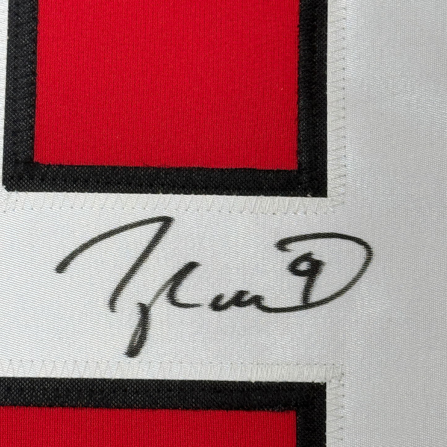 Close-up of handwritten signature on white fabric strip between red and black sections of jersey.