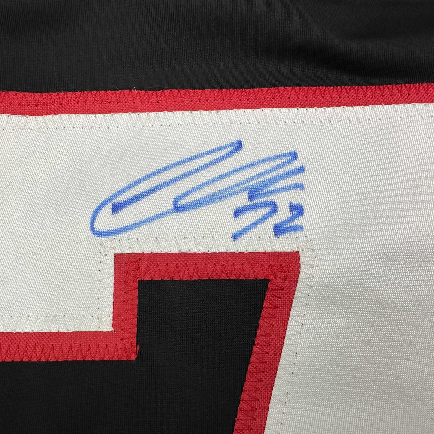 Close-up of blue autographed signature on white fabric panel with red and black striped hockey jersey border.