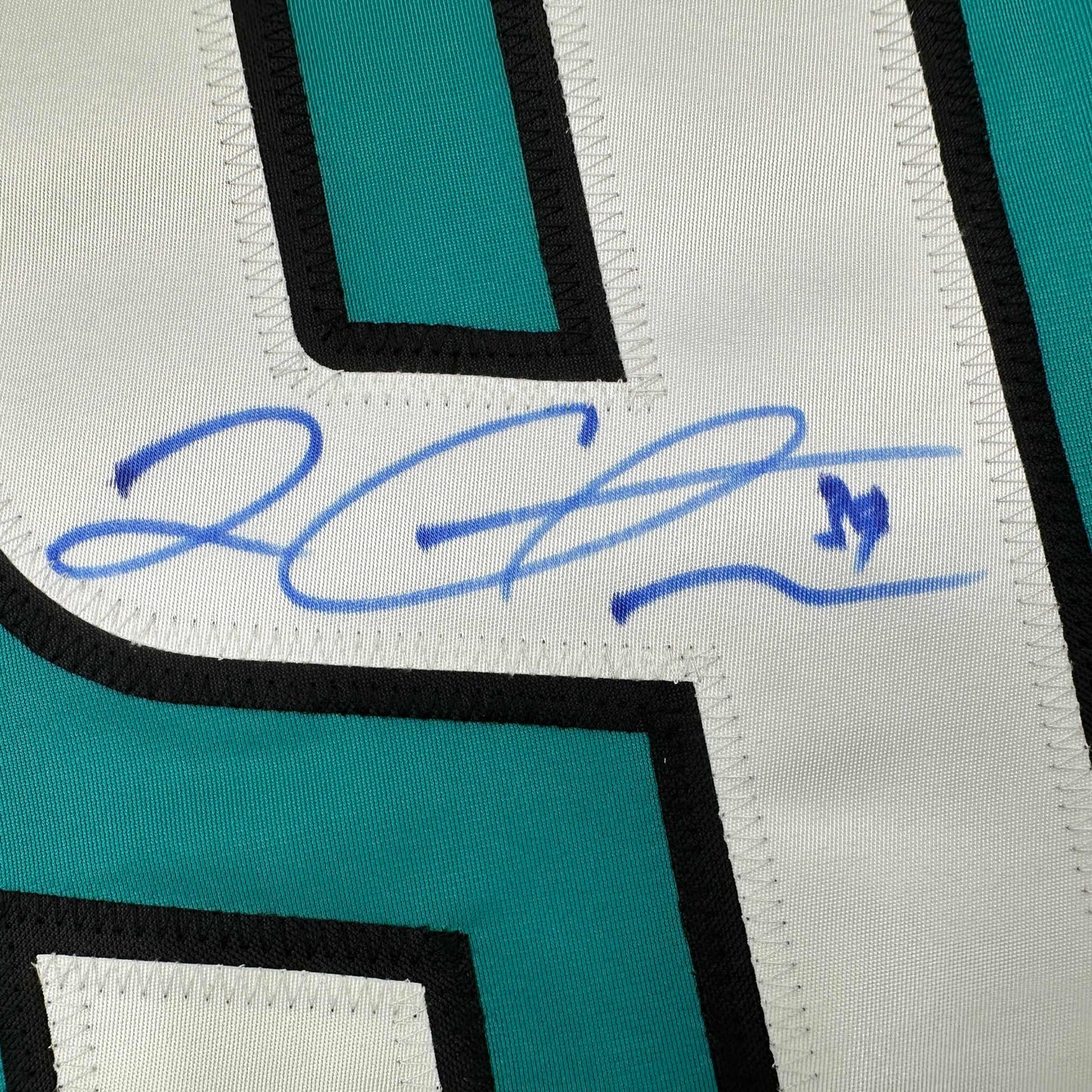 Close-up of blue autograph signature on white fabric panel with teal and black striped jersey trim visible.