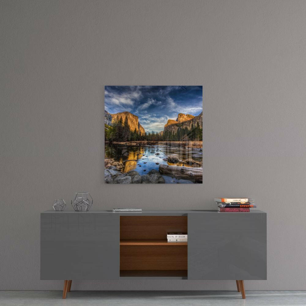 Square glass wall art featuring a mountain valley landscape with golden cliffs, forested slopes, reflective river, and blue sky, displayed above a mid-century modern console table.