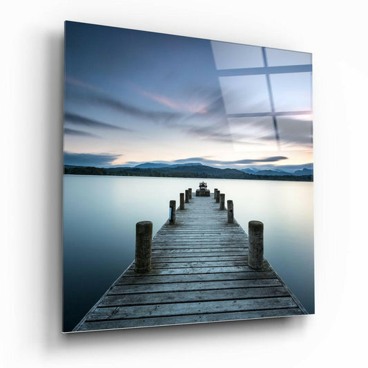 Wooden jetty extending into calm blue lake at dusk with mountains and sky reflected in glass panel overlay.