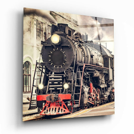 Front-facing view of a black steam locomotive with red cowcatcher and illuminated headlights against an industrial brick building backdrop.