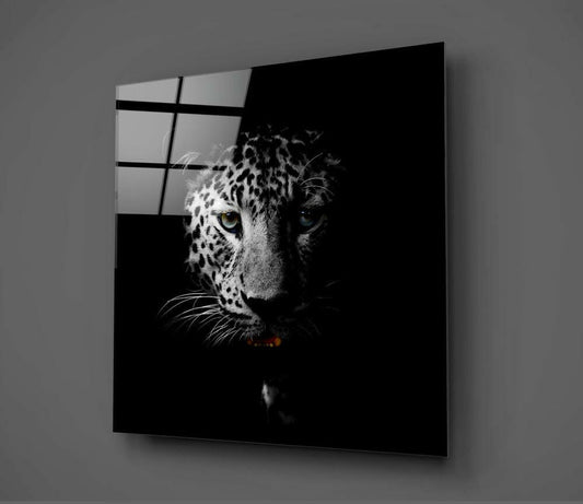 Glass wall art mounted on gray wall displaying a leopard's face emerging from black background, with window reflection visible in upper left corner.