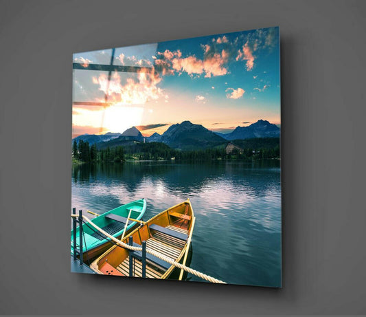 Green and yellow canoes tied to a dock on a serene alpine lake at sunset, with snow-capped mountains and forest silhouetted against an orange and blue sky, displayed as glass wall art.