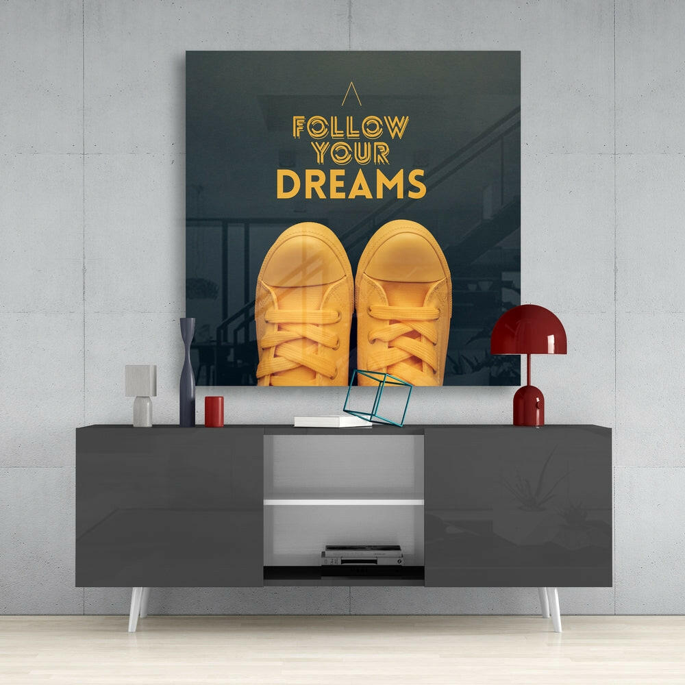 Glass wall art displaying yellow sneakers and gold "Follow Your Dreams" text mounted above a dark gray media console in a modern interior setting.