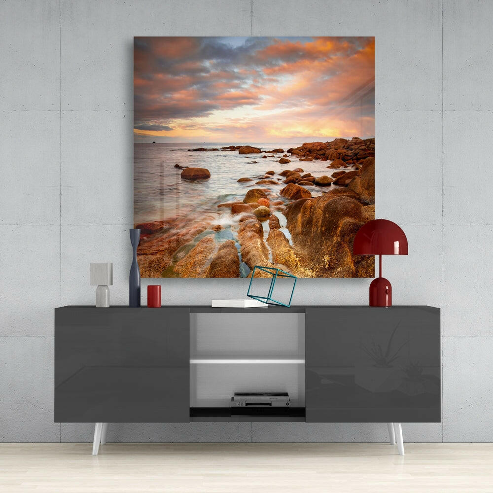 Glass wall art depicting a rocky coastline at sunset with golden and orange clouds displayed above a dark gray media console.