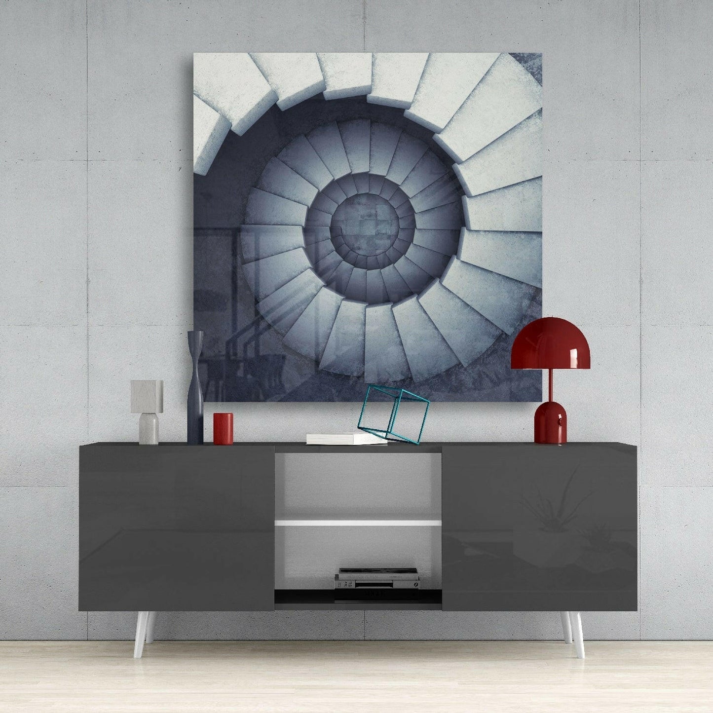 Spiral staircase wall art displayed above a dark gray media console with red lamp and accessories in minimalist interior setting.