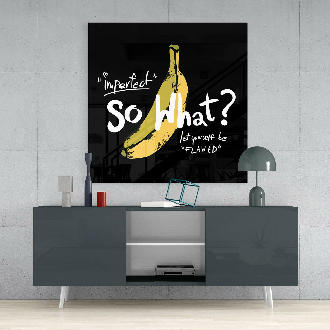 Black glass wall art with yellow banana illustration and white text reading "Imperfect" So What? let yourself be "FLAWED" displayed above gray media console.
