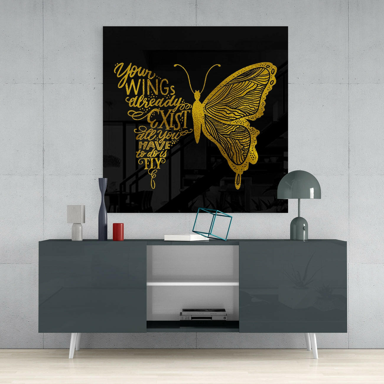Gold butterfly and script text on black glass art displayed above gray media console with white legs and decorative accessories.