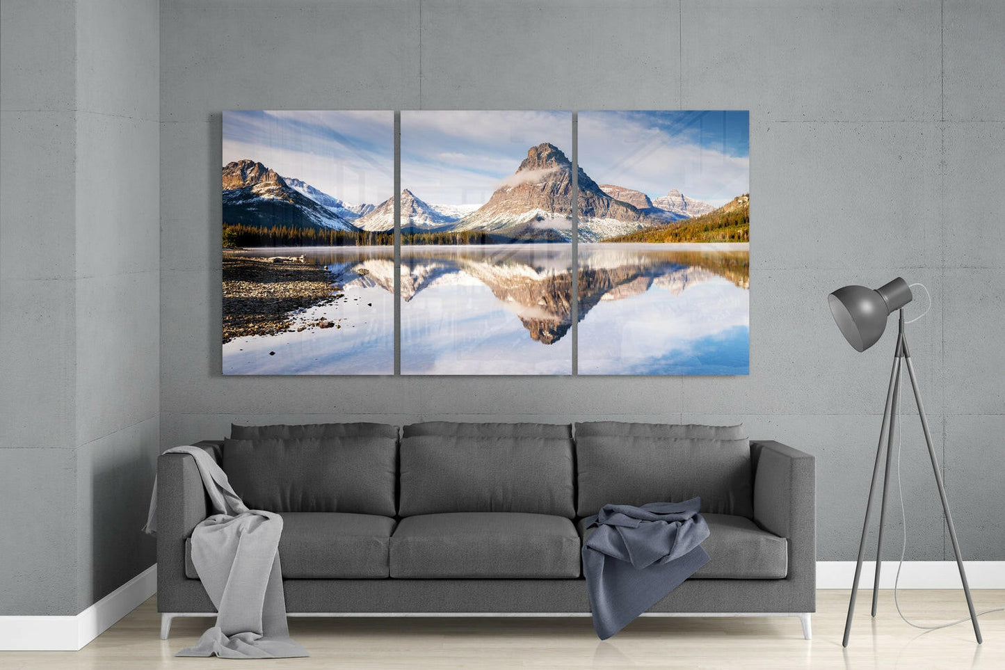 Three-panel glass wall art displaying mountain lake reflection with snow-capped peaks, displayed above gray sofa in modern living room.