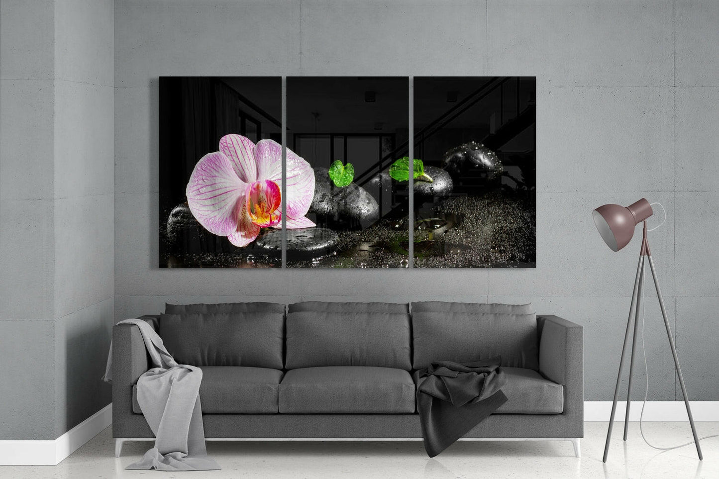 Three-panel glass wall art displaying pink orchid flower and black stones with green foliage against grey living room wall above sofa.
