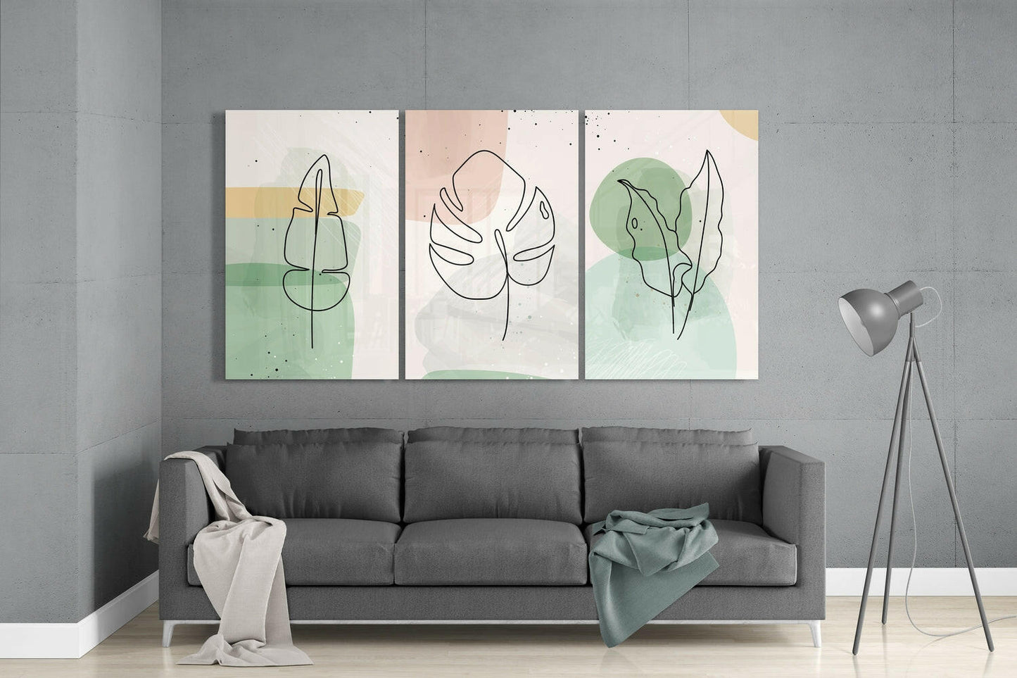 Three glass wall art panels with minimalist line-drawn leaves and watercolor washes in green, beige, and pink tones displayed above a gray sofa.