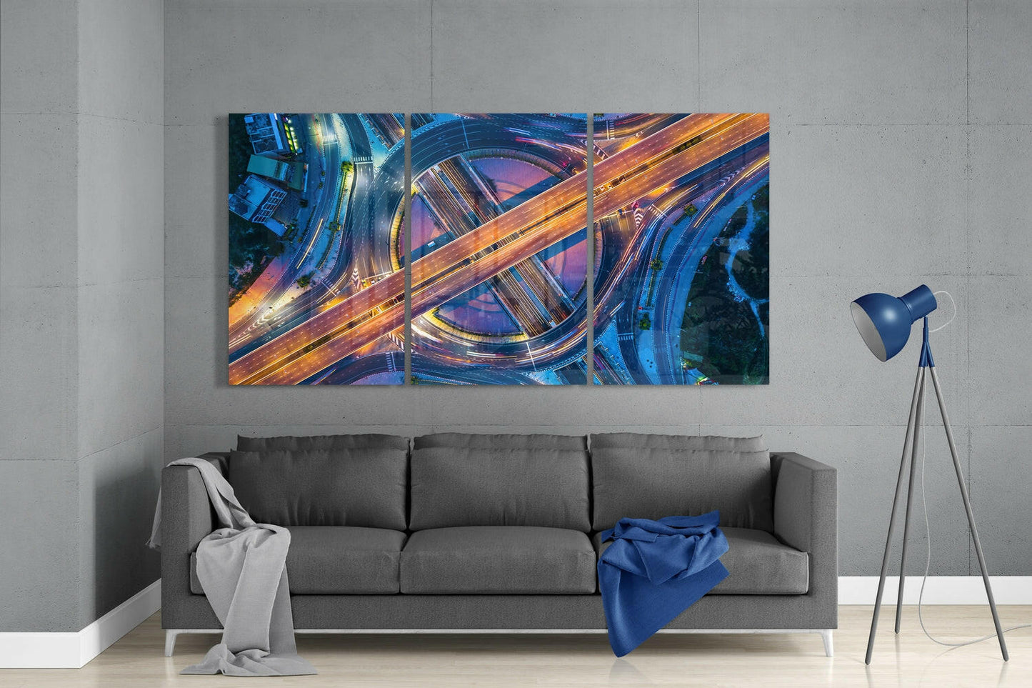 Triptych glass wall art depicting illuminated aerial highway interchange in blue and orange tones, installed above gray sofa in modern living room.