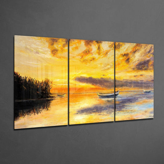 Three-panel glass wall art displaying a golden sunset over water with moored boats and dark shoreline vegetation against gray wall.