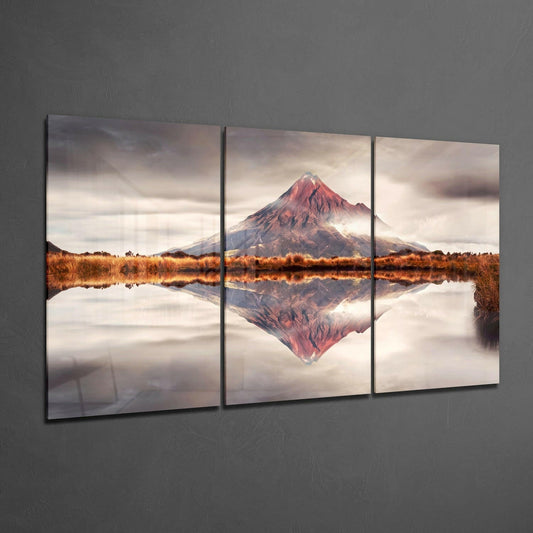 Three-panel glass wall art displaying a snow-capped red volcano reflected in still water with golden marshland, mounted on gray wall.