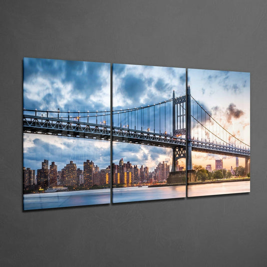 Three-panel glass wall art displaying a suspension bridge spanning a river with illuminated city skyline at dusk mounted on gray wall.