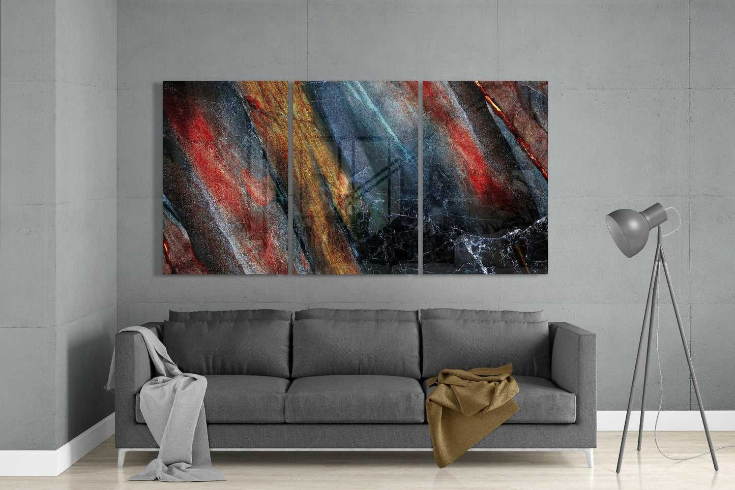 Three-panel abstract glass art with diagonal stripes in red, gold, blue, and black hung above a gray sofa in a modern interior.