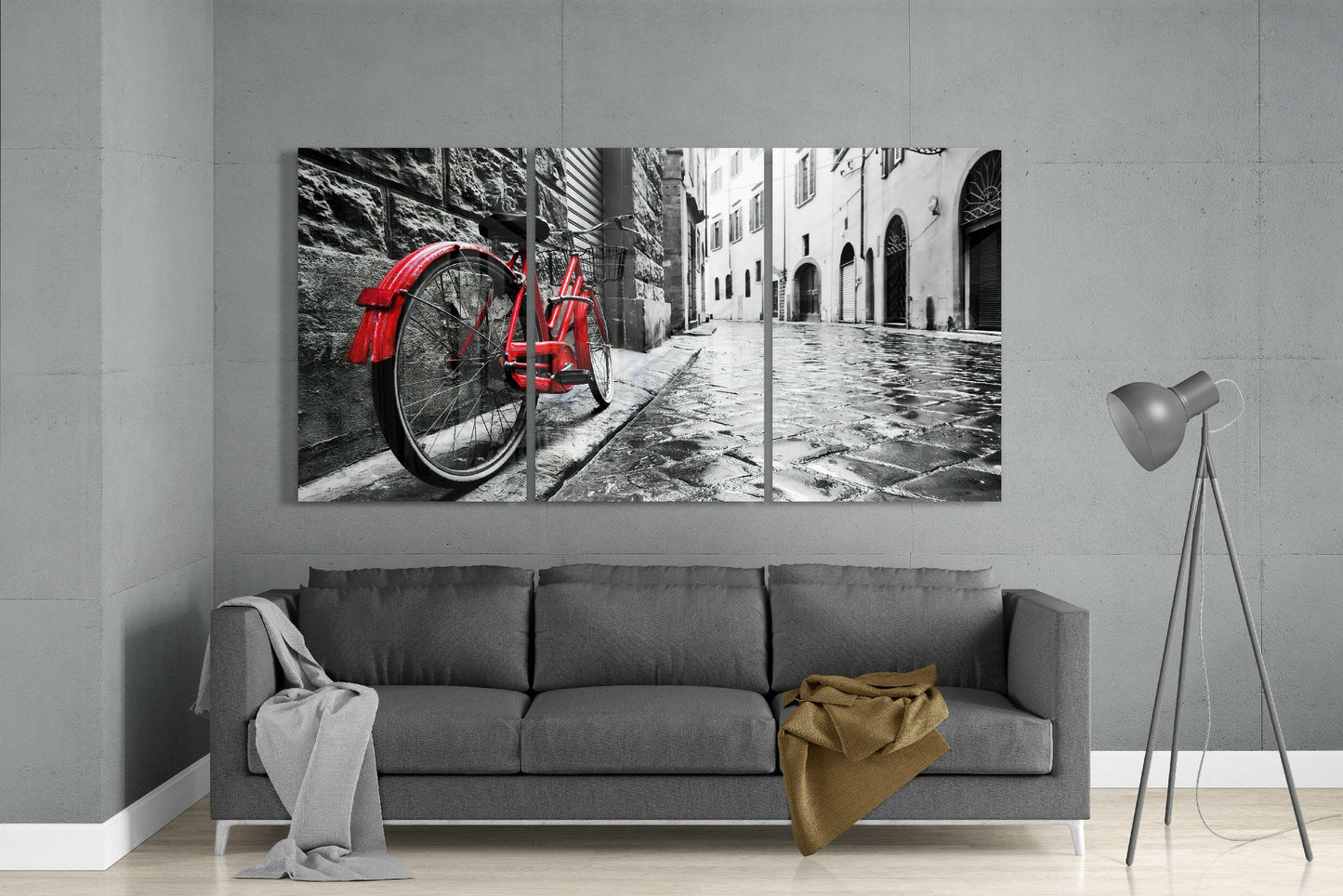 Three-panel glass art mounted above gray sofa displaying red bicycle on cobblestone street with monochrome buildings.
