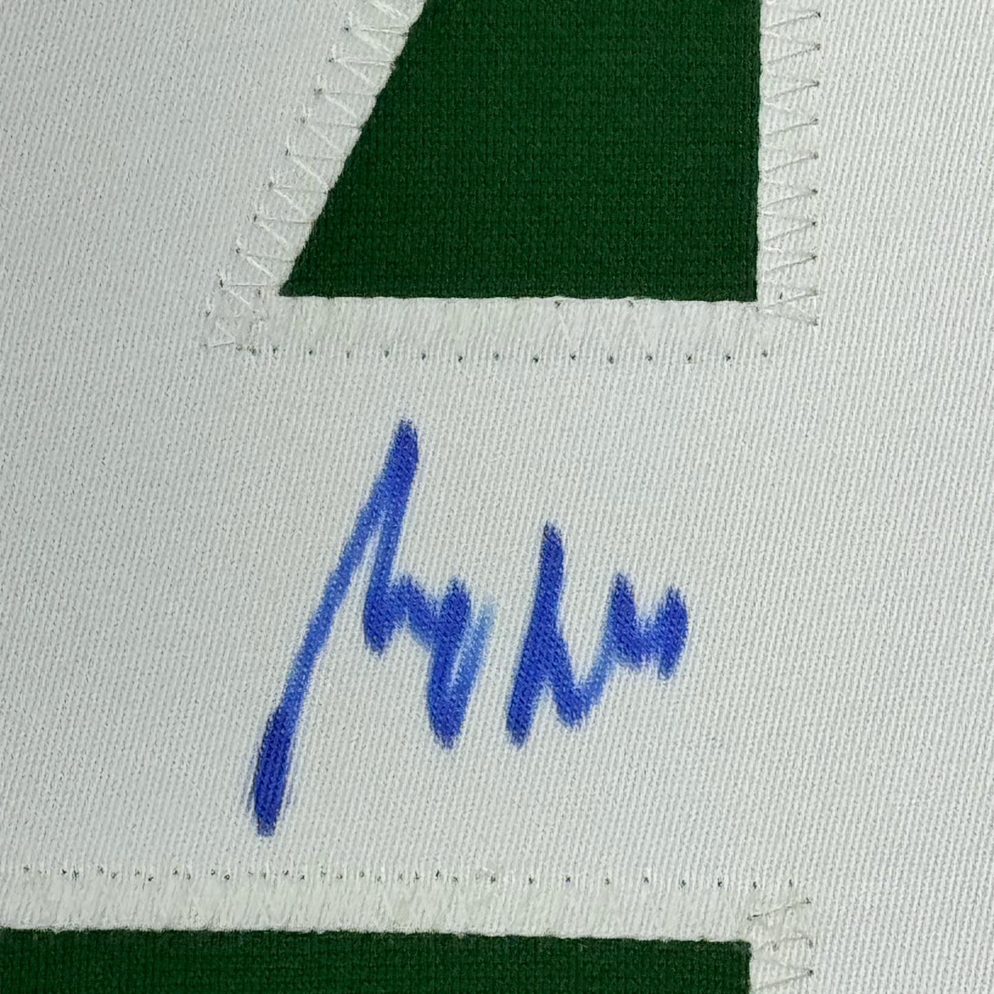 Close-up of blue autograph signature on white fabric with green jersey sections visible at top and bottom.