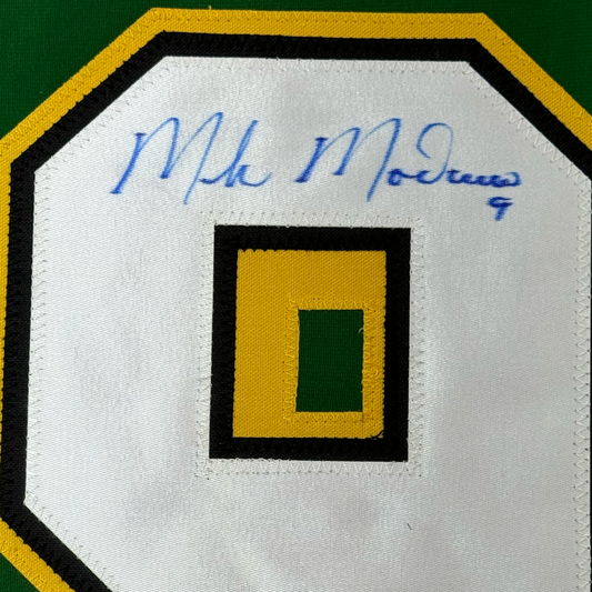 Close-up of white fabric label with blue handwritten signature and concentric square design in yellow, black, and green.