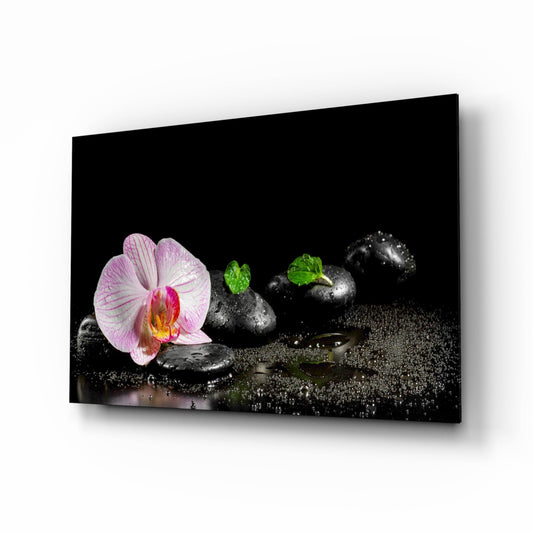 Glass wall art displayed at an angle showing a pink orchid flower, black zen stones, green moss, and water droplets on black sand against a dark background.