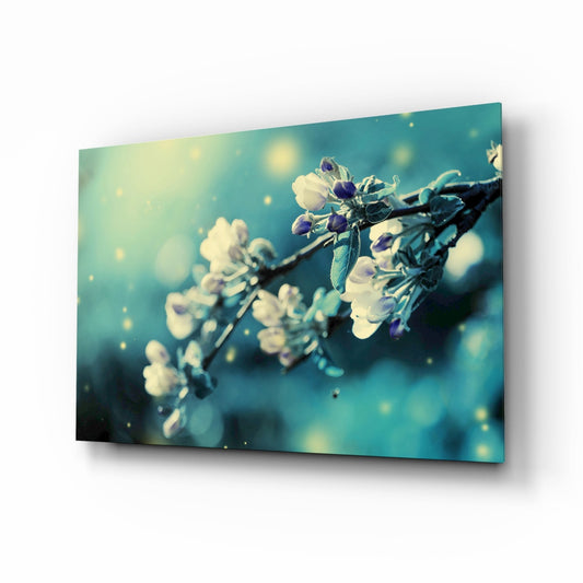 Glass wall art panel displaying a frost-covered flowering branch against a soft teal and cream bokeh background with warm light sparkles.