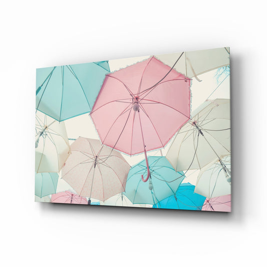 Angled product shot of a glass wall art panel displaying overlapping pastel pink, turquoise, cream, and blue umbrellas viewed from above.