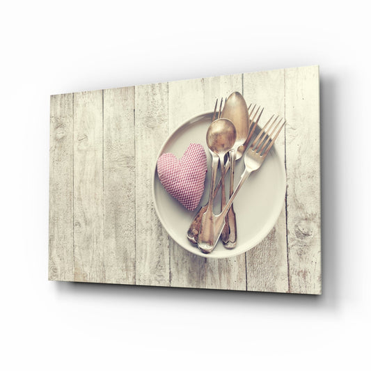 Angled view of glass wall art showing white plate with pink gingham heart and vintage cutlery on whitewashed wooden background.