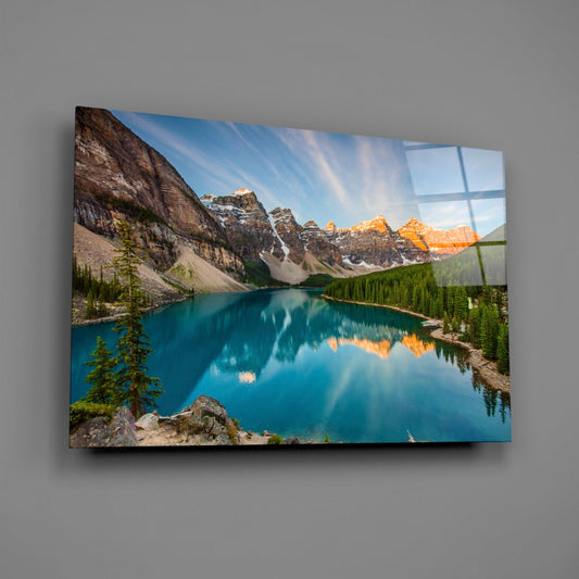 Angled view of glass wall art mounted on gray wall, displaying turquoise alpine lake with snow-capped mountains and evergreen forests at sunrise.