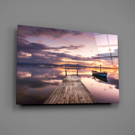 Wooden jetty extending into calm violet-toned water at sunset with moored boat and mountains in distance, displayed as glass wall art.