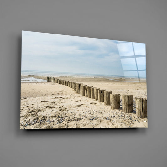 Wooden breakwater posts receding into distance across sandy beach with cloudy sky, mounted on gray wall.