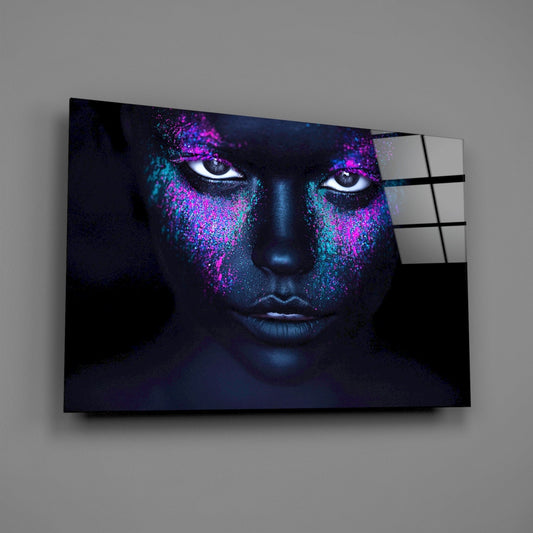 Glass wall art panel displaying a dark-skinned face with magenta and cyan glitter makeup effect, mounted on gray wall at angle.