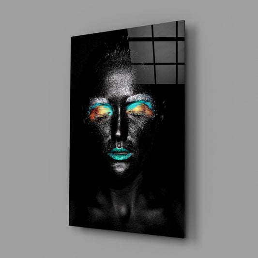 Glass wall art panel mounted on gray wall showing portrait of person with teal lips and gradient eyeshadow in turquoise, gold, and orange tones against black background.