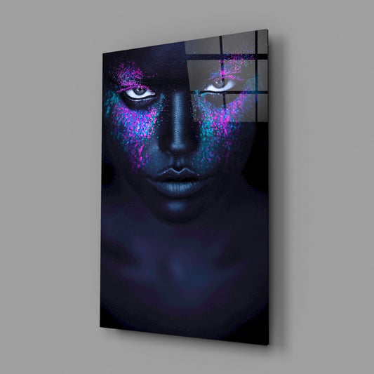 Angled view of glass wall art displaying a dark-skinned face with glowing pink and turquoise powder effects around the eyes against a black background.