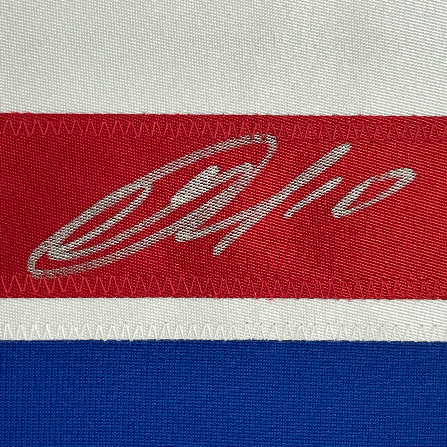 Close-up of embroidered signature on red stripe of hockey jersey with white and blue stripes visible.