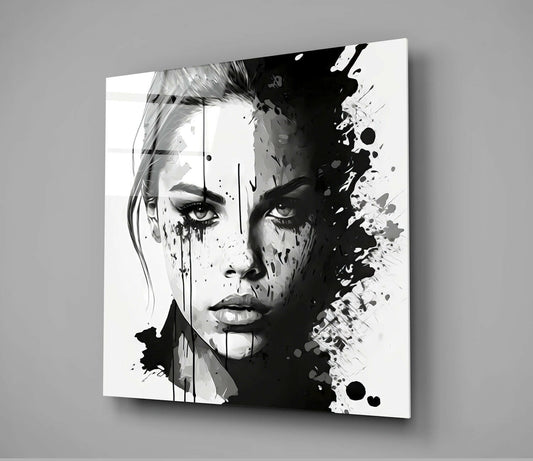 Black and white glass wall art featuring a woman's face with ink splatter effects, displayed at an angle against a gray wall.