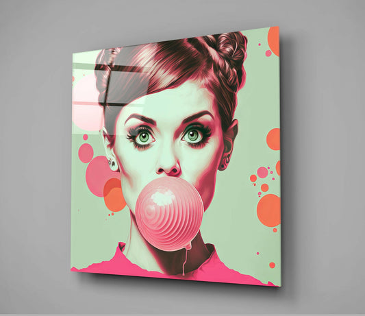 Angled view of glass wall art showing a retro-styled woman's face blowing a large pink bubble against a mint green background with coral circular accents.